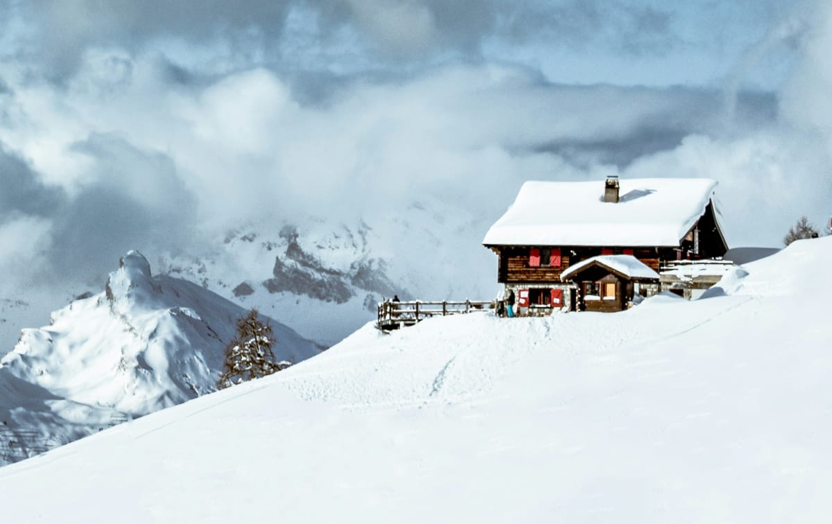 A snow covered house perched on a mountain. Large clouds swarm above it, and more mountains are to the side of it.