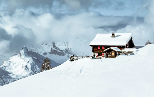 A snow covered house perched on a mountain. Large clouds swarm above it, and more mountains are to the side of it.