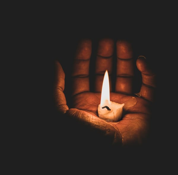 A dark skinned hand, cradling a candle nub in their palm. Wax is trailing between the fingers as the flame stands high.